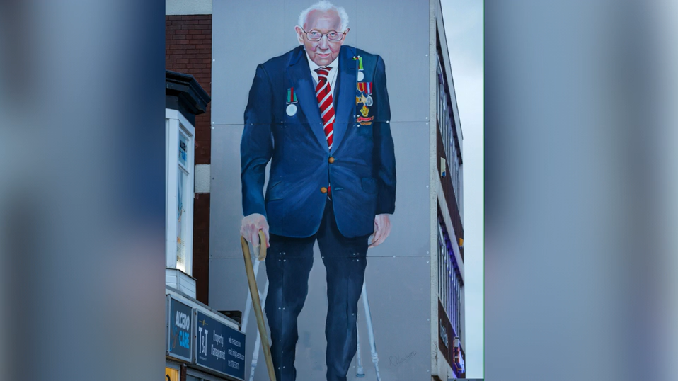 Robert Newbiggin's painting of Captain Tom Moore on the side of an office building in Southport. Captain Tom is wearing a blazer with his medals on and is leaning on his walking stick. 