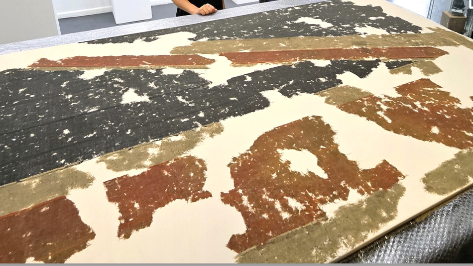 The damaged flag is laid out on a table. Material is missing in several areas and there are gaps and deterioration in what cloth has survived. It is red, navy blue and the white is a dirty, aged yellow. On the right there are two red and white diagonal stripes in the design.