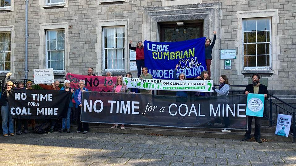 A group of campaigners standing outside a stone building. They're holding signs and banners which read 'No time for a coal mile', 'Leave Cumbrian coal underground' and 'South Lakes on climate change action'.