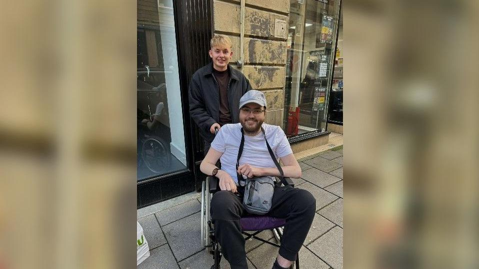 Stephen Jefferson and his brother-in-law Daniel Swales smiling at the camera. Mr Jefferson has blonde hair is dressed in black trousers, jacket and t-shirt. Mr Swales has a cap on and is wearing black trousers, a white t-shirt and a grey cross body bag.