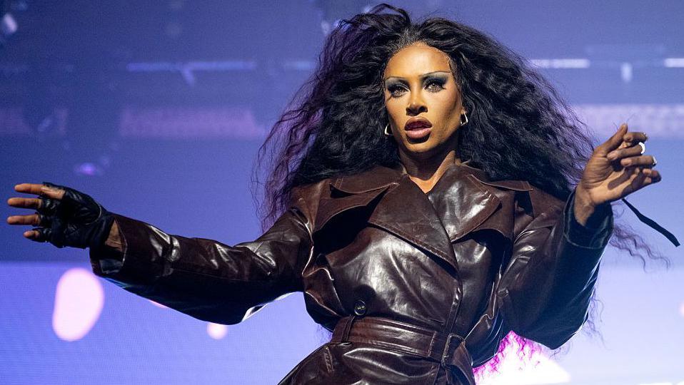 Drag performer Tayce is on-stage at Manchester Pride in 2025. She has long black hair and is wearing a brown leather coat which is fastened at the waist with a thick belt. Her arms and hands are outstretched towards the audience and she is stood in front of bright purple lighting.