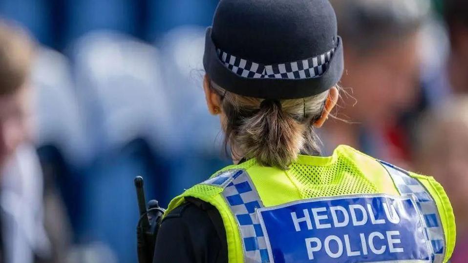 A female police officer wearing a hat and uniform faces away from the camera. Her uniform reads: HEDDLU POLICE