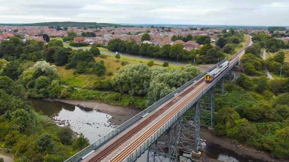 A two-carriage passenger train is crossing a steel railway viaduct over a shallow river, with a town in the background 