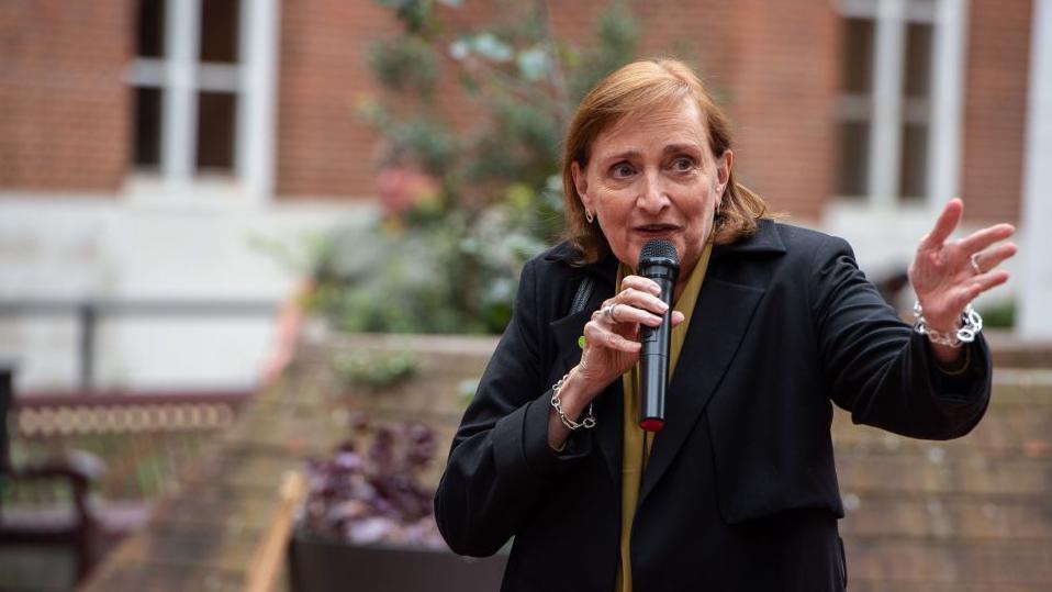 Emma Dent Coad, a woman with short red hair wearing a black coat and yellow scarf, speaks into a microphone and stands in front of red-brick houses.