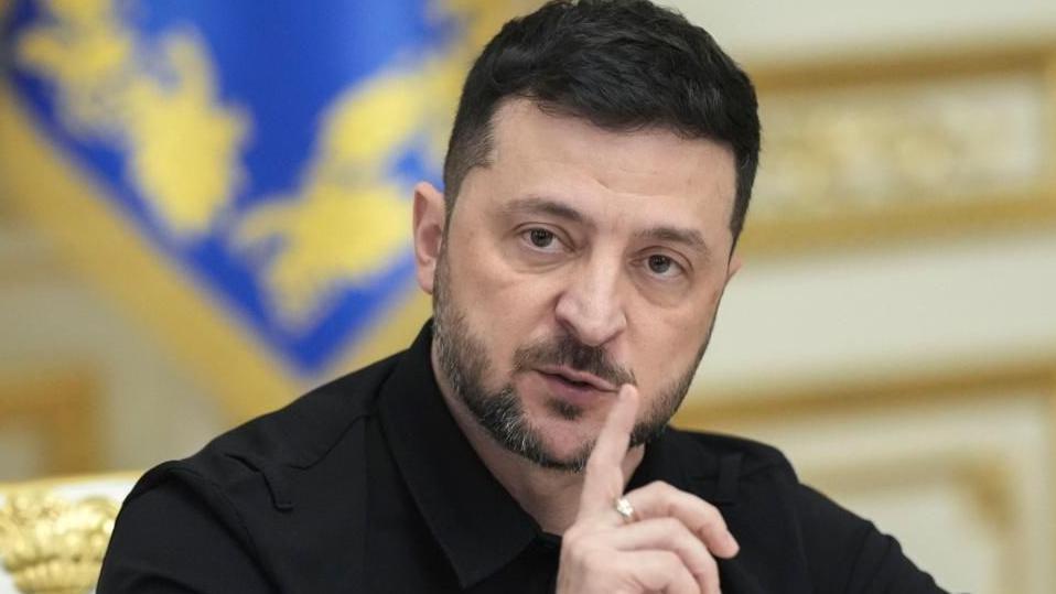 A head and shoulders image of Volodymyr Zelensky wearing his usual black suit and holding his index finger near his mouth. He has a near  beard and moustache.