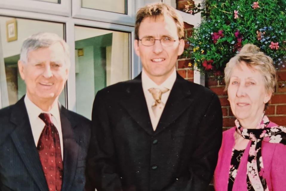 Ray and Simon Stokes, both wearing shirts, ties and jackets, smiling, in front of a window, next to Jill Stokes wearing a pink jacket, with brickwork and flowers behind her.