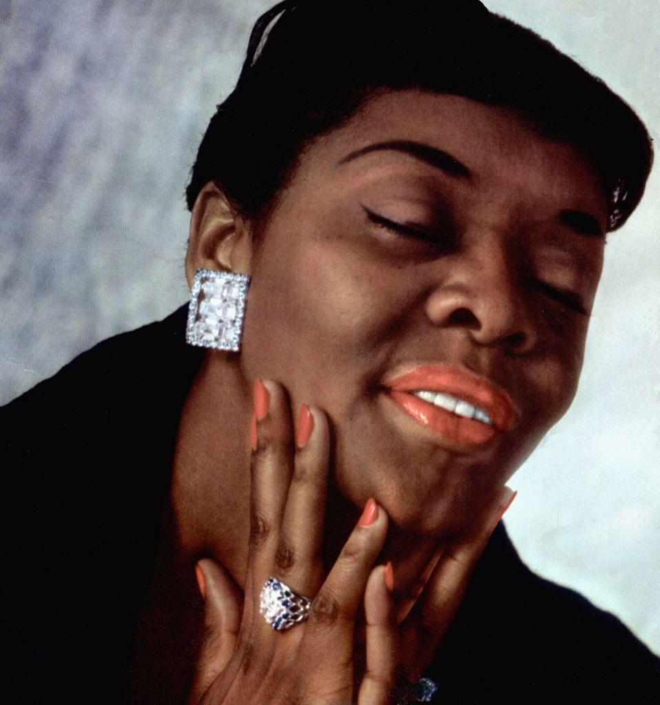 Dinah Washington with her hand on her chin and her eyes closed. She is wearing large square diamond earrings and a diamond ring.