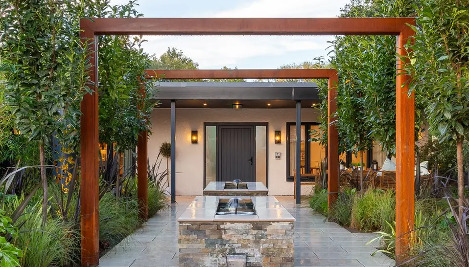 Welcoming entry to a charity that supports people with their mental health. This picture shows an orange trellis, surrounded by plants and small fountains are running which are surrounded by stone and bricks.