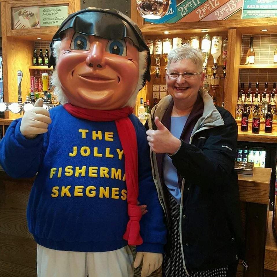 Mascot with black hat, white hair, blue jumper with the Jolly Fisherman Skegness written on it, red scarf, white trousers doing a thumbs up sign with one hand. A lady with short light hair and glasses smiling in a blue top and light coat also doing a thumbs up sign with one hand. They are stood in a bar.