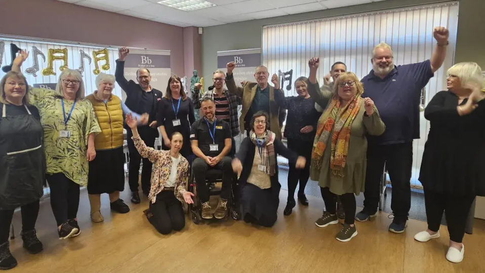 In a room with a wooden floor, fifteen people, including a man in a wheelchair, raise their arms in celebration
