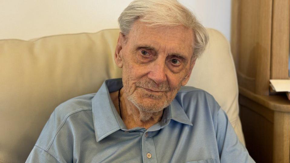 A man with white and grey hair is wearing a blue shirt. He is sitting on a sofa.