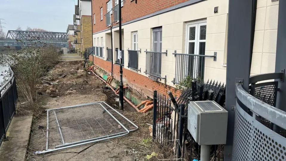 An uneven footpath between the River Nene and a block of flats- with a metal grill covering the sinkhole.