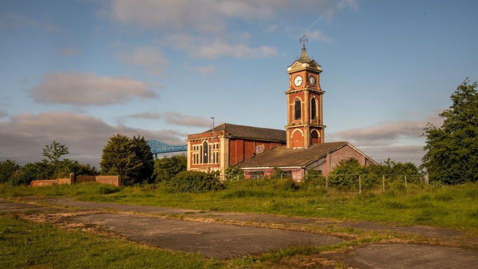 Middlesbrough's derelict Old Town Hall in the Middlehaven area. It is built from red brick and has a tower with a spire. It is surrounded by wasteland. The Tees Transporter Bridge can be seen in the distance.