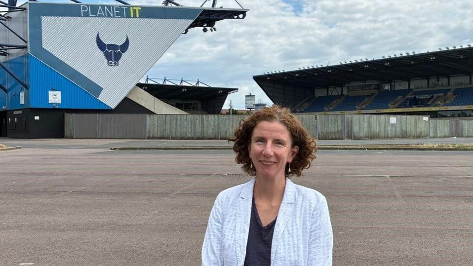 Anneliese Dodds smiling outside near the current home of Oxford United FC, the Kassam Stadium on a cloudy day. The club's bull logo can be seen.