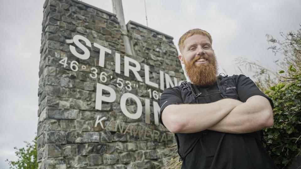 Russ Cook standing in front of a wall. He is wearing a black t-shirt and has his arms crossed. He is smiling and looking at the camera. 