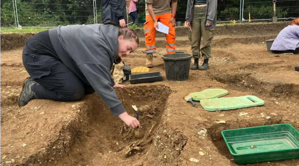 Thalia Argrave is wearing a grey hoodie and black trousers, and has brown and pink hair in a plait. She's kneeling on the floor and leaning into a pit to brush soil away from a partially-excavated skeleton. There are buckets and tools on the ground around her