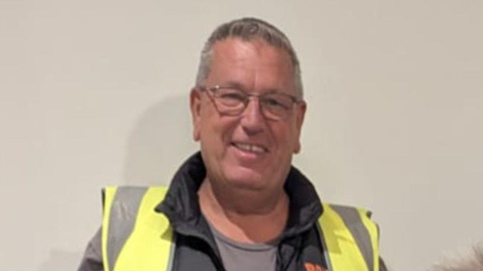 Tony Steel with short grey hair and glasses is standing against a grey wall. He is wearing a fluorescent hi-vis jacket.