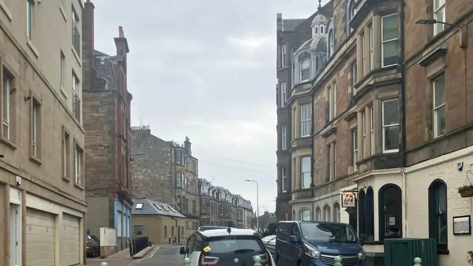 Bath Street in Portobello. Light-stone tenement buildings can be seen on either side. A white and black car is in the centre of the foreground. A dark-coloured van is parked on the right side of the road.