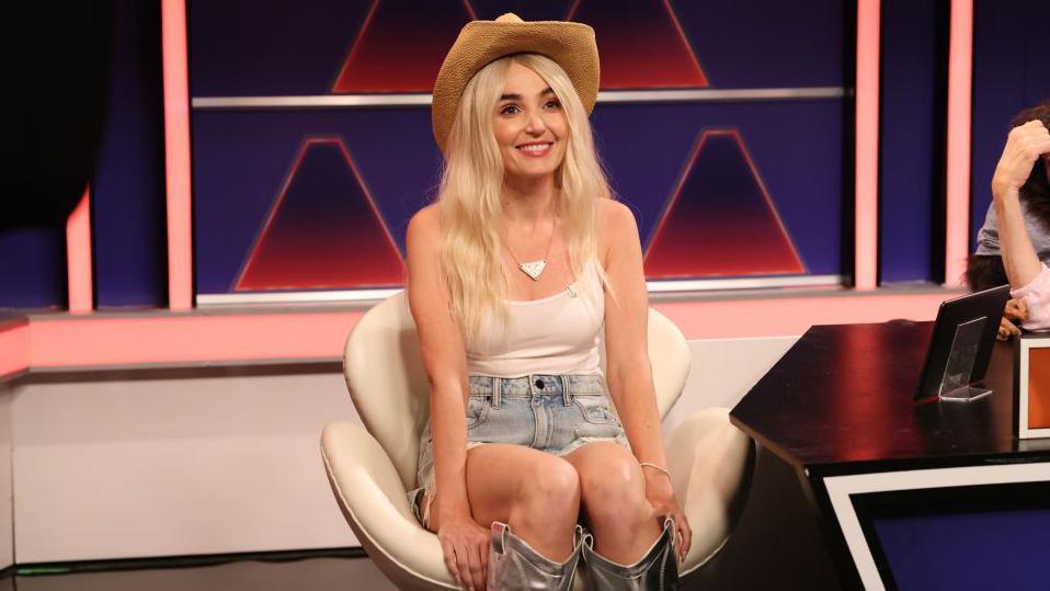 Actress Chloe Fineman sits in a TV set on a white chair, wearing a cowboy hat, white vest top, denim shorts and silver cowboy boots. She is impersonating Hawk Tuah girl Haliey Welch. 