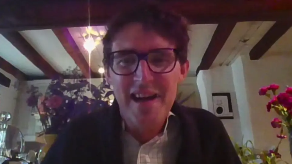 Simon Boas, who has brown hairm is man wearing glasses, a checked white shirt and a navy cardigan sitting facing the computer camera in a living room.