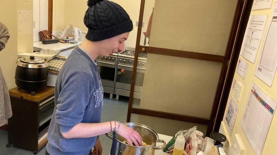 Aimee Clasby, trainee youth development worker at a baking session. She wears a blue sweatshirt and a black bobble hat and is mixing some food in a saucepan.