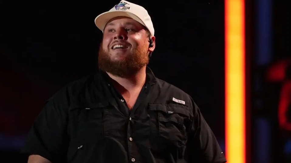 Luke Combs is wearing dark shirt and a cap on stage and is smiling. 