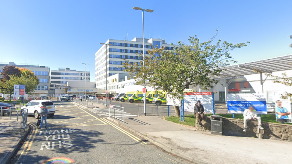 The entrance to the hospital in Barnsley. Two people can be seen sitting on a wall on the right, next to a bin. The words "Thank you NHS" can be seen on the road leading to the hospital entrance. Several ambulances are parked outside the building.