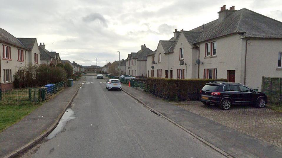 A street with two-storey residential buildings on either sides. There are four cars parked on driveways and on the road.