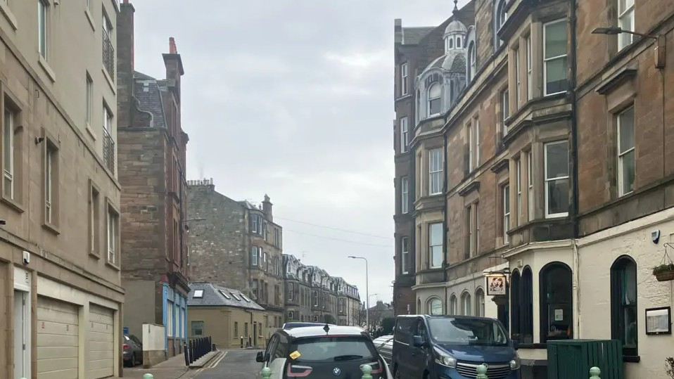 Bath Street in Portobello. Light-stone tenement buildings can be seen on either side. A white and black car is in the centre of the foreground. A dark-coloured van is parked on the right side of the road.