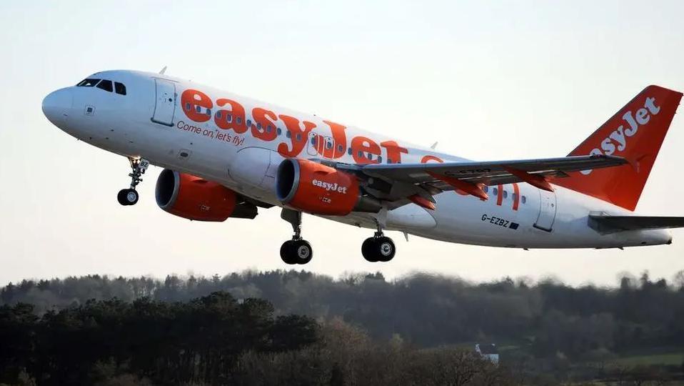 An orange and white EasyJet plane taking off from a runway. It is in the air with its wheels still down and there are trees below.