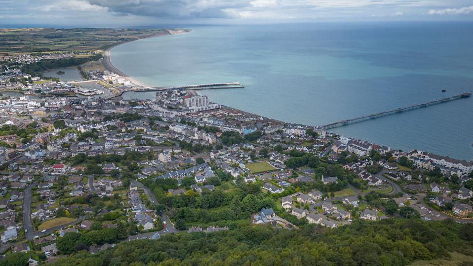 An aerial view of Ramsey showing the roads and houses of the town, and the curve of the bay, which has two piers extending out to sea.