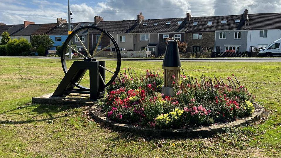 View of a flower bed centred around a sculpture of a miner's lamp and another of the wheel of a pit head. A row of terraced houses are in the background.