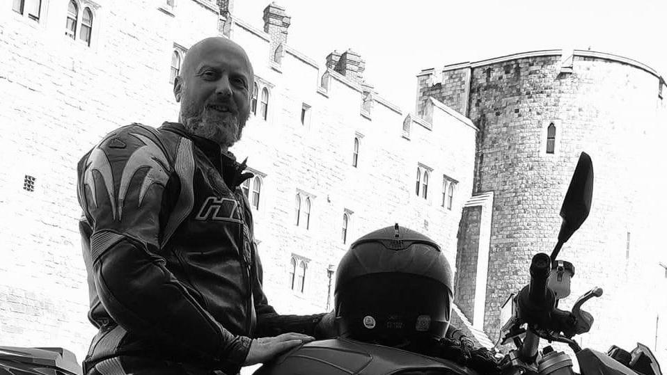 Pawel Dlugosz is sat on a motorcycle in front of a castle. The photo is in black and white. He is bald and has a beard, and is wearing a leather jacket and trousers.