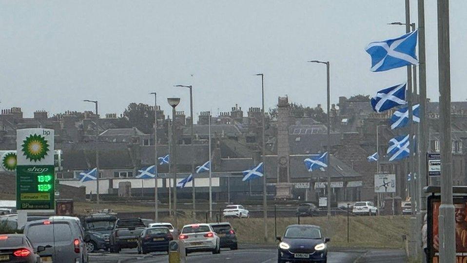 A row of blue and white saltire flags attached to lampposts on a busy road in Peterhead. Several cars are passing by.