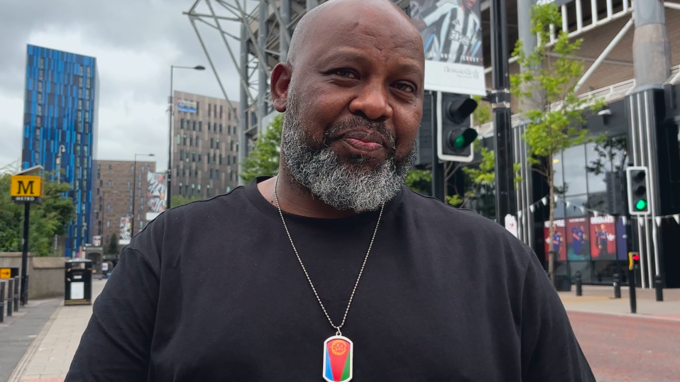 Mehari Estifanos, a shaven-headed man with a black and grey beard, is seen wearing a black t-shirt and a necklace with a pendant of an Eritrean flag outside Newcastle United's stadium