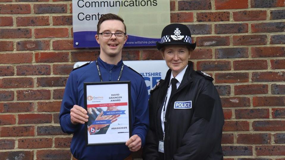 Man in blue shirt holding a frame which says 'David Grainger Award' stood next to a woman in a black jacket which says 'police' in blue and wearing a black hat. They are stood in front of a brick wall.