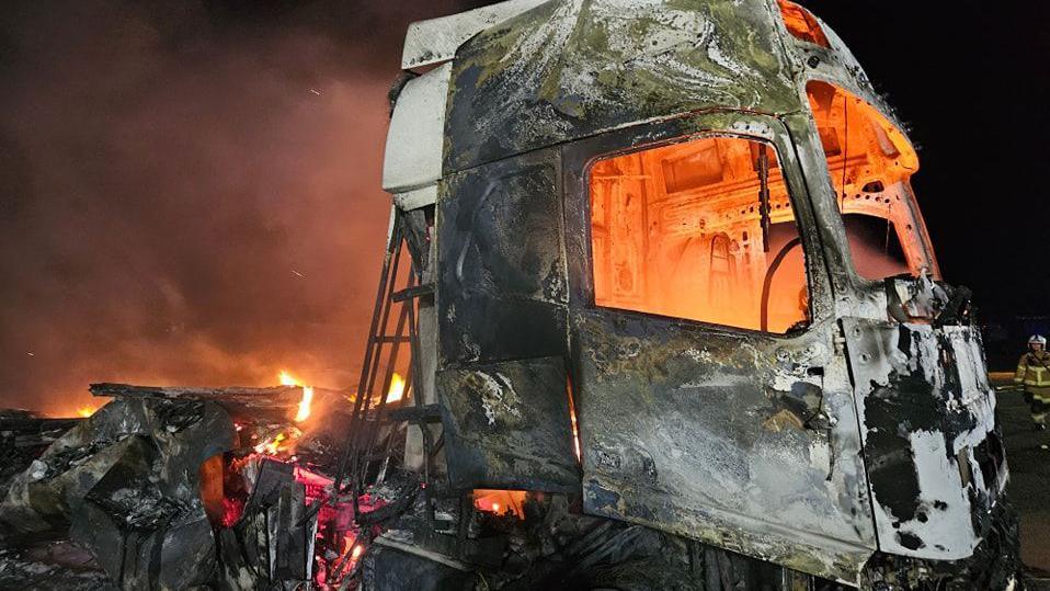 A charred lorry that has been almost entirely consumed by flames. The metal is red hot and the cab is glowing orange from flames 