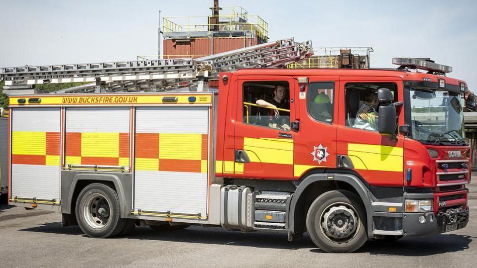Red and yellow fire engine with ladder on roof. The web address WWW.BUCKSFIRE.GOV.UK is visible in red lettering above the side doors. Fire fighters can be seen in the cab.