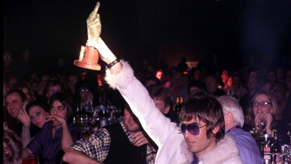 A photo of Liam Gallagher at an NME award event, holding up a trophy in the shape of a hand giving a middle-finger salute