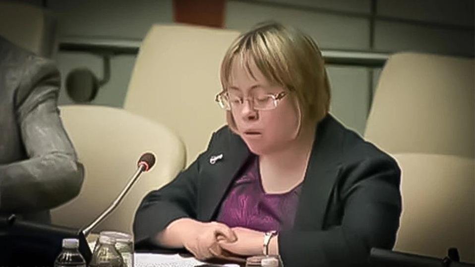 Sara Pickard is wearing a purple top and a black blazer. She is speaking into a microphone at the United Nations while reading from a piece of paper. She has a blond/brown bob and wears glasses.