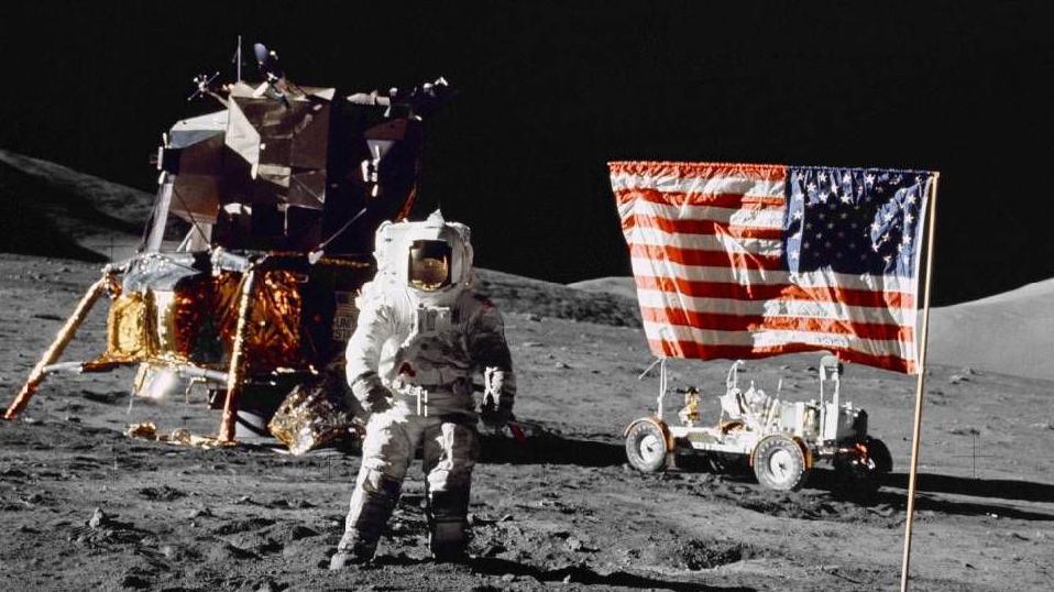 A picture of an astronaut on the Moon. Next to him is a United States flag, and behind him are devices used on the Moon. 
