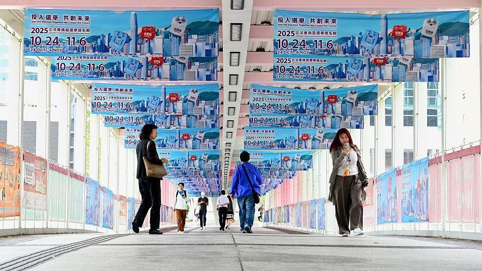Hongkongers walk on an overhead bridge festooned with blue posters advertising the 2025 Hong Kong Legco election campaign