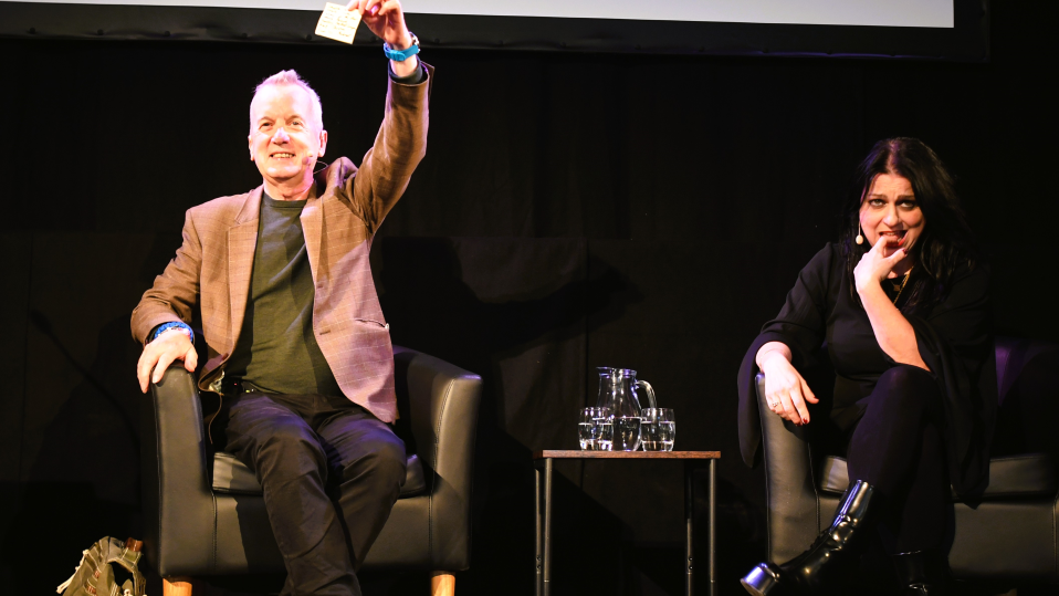 Frank Skinner and Janina Ramirez in armchairs on stage. Frank Skinner holds up a piece of paper, while Janina rests her head on her hand. A small table with a glass pitcher and glasses sits between them, set against a dark indoor backdrop.