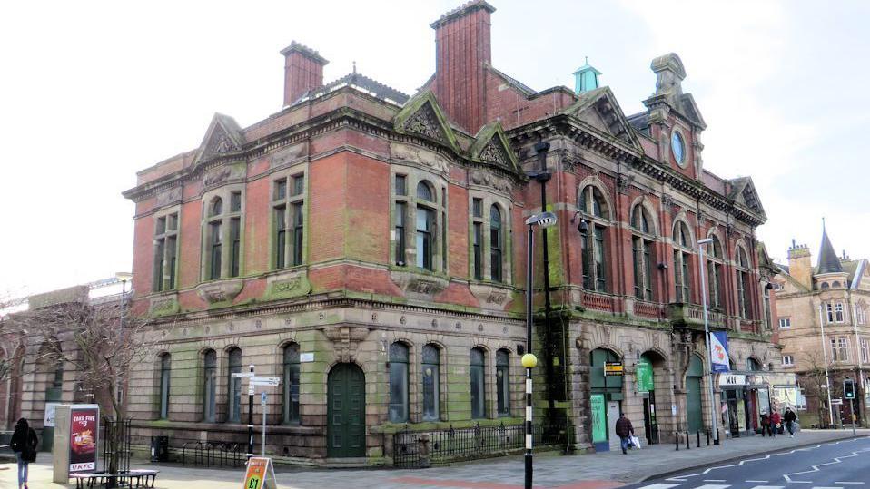 A large old indoor market building. People can be seen walking past the front entrance on the pavement. A road with a pedestrian crossing is visible on the right.