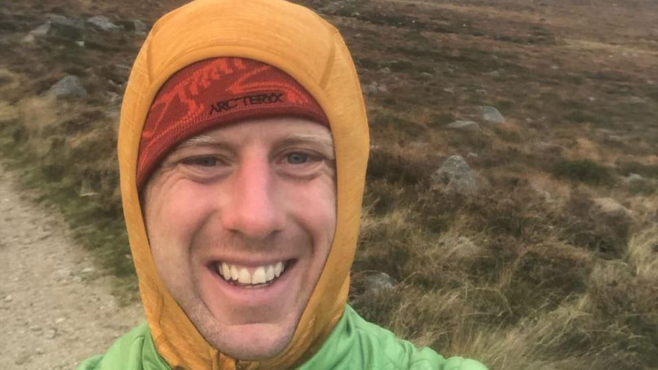 Andrew McMurray is wearing a lime green jacket, he has a dark yellow hood or snood pulled over his head and a dark red hat underneath that. Behind him moorland can be seen.