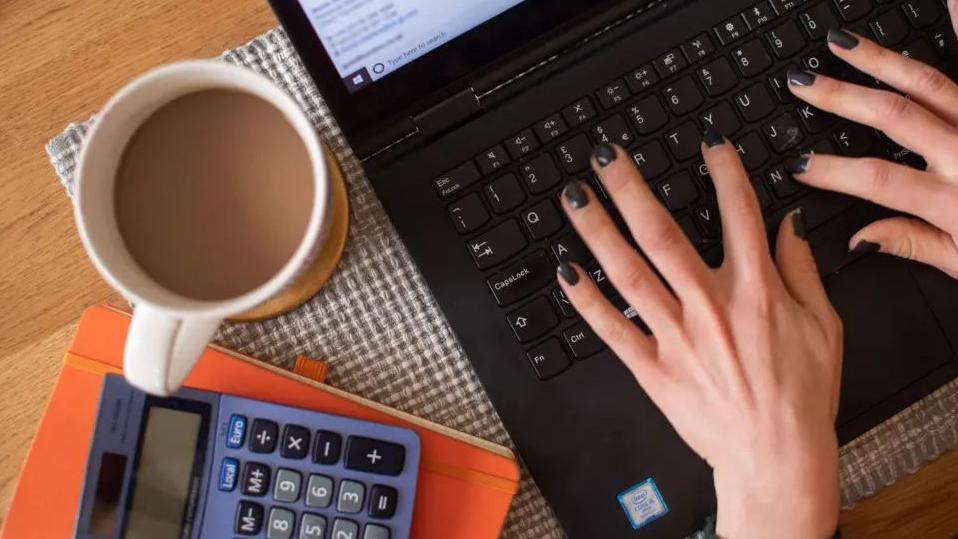 A view of a woman's hands typing on a black laptop. There is a cup of tea in a white mug next to the device along with a calculator on top of an orange notebook.