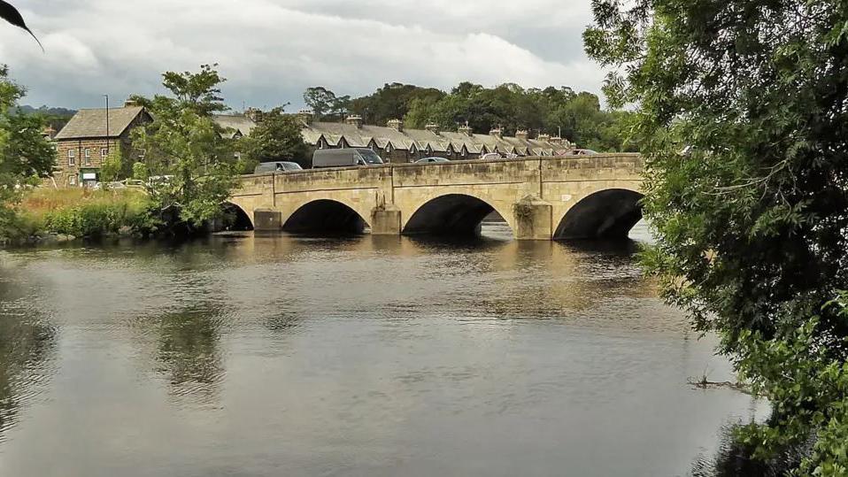 The existing bridge over the Wharfe in Otley is a seven-span stone bridge which dates back to the 13th Century.  It is shown here with houses in the background and trees on the river bank.