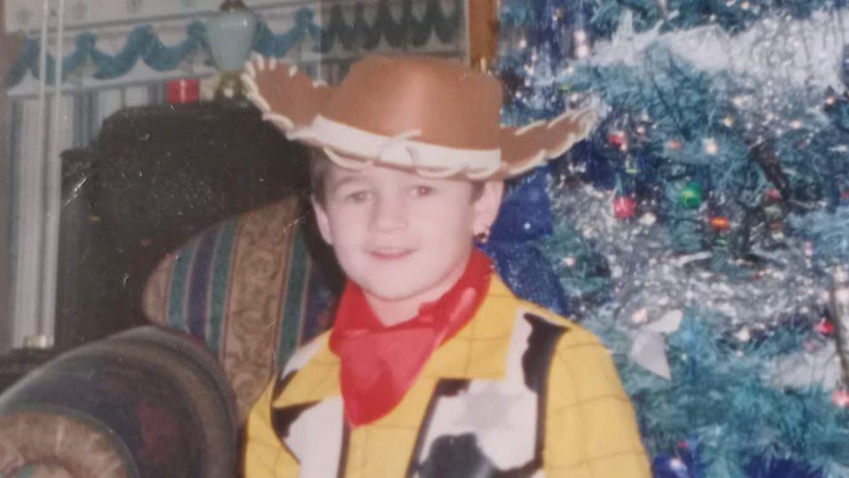 A photograph of Kaine Fletcher as a child, wearing a Woody costume from Toy Story', standing in front of a Christmas tree
