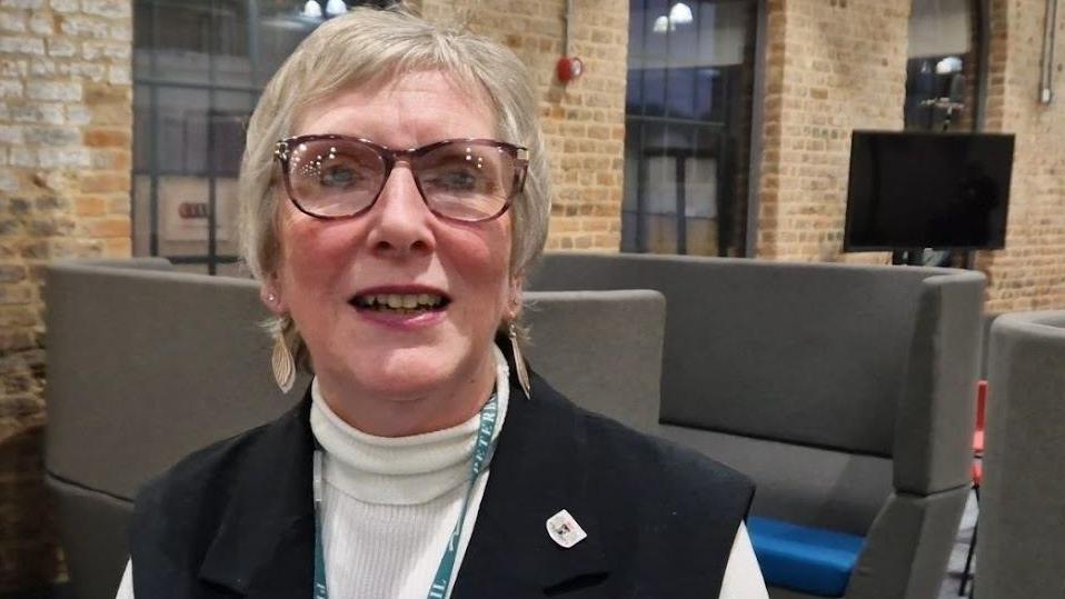 Alison Jones has short fair hair and wears glasses. She is wearing dangly earrings, a cream polo neck jumper and sleeveless black jacket. She is sitting down near banks of soft seating and in a corporate room with exposed bricks and large windows.