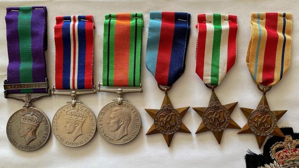 Three circular, silver medals with colourful ribbons, laid on a white cloth alongside three bronze medals shaped like stars with colourful ribbons.